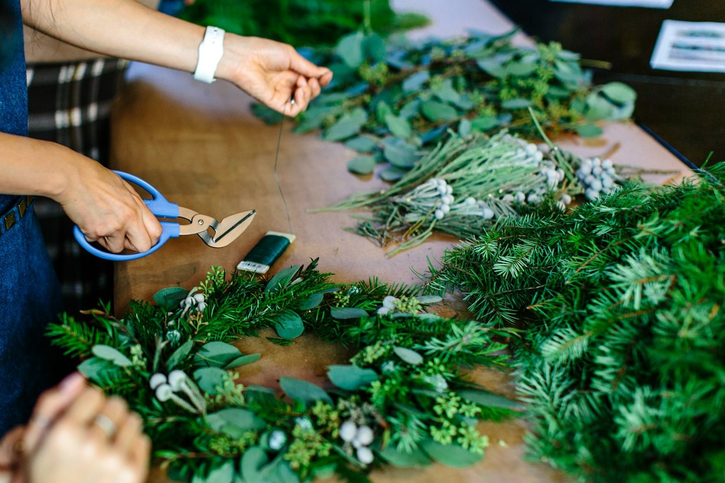 Hands at a wreath-making workshop table covered with greenery, shears and wire