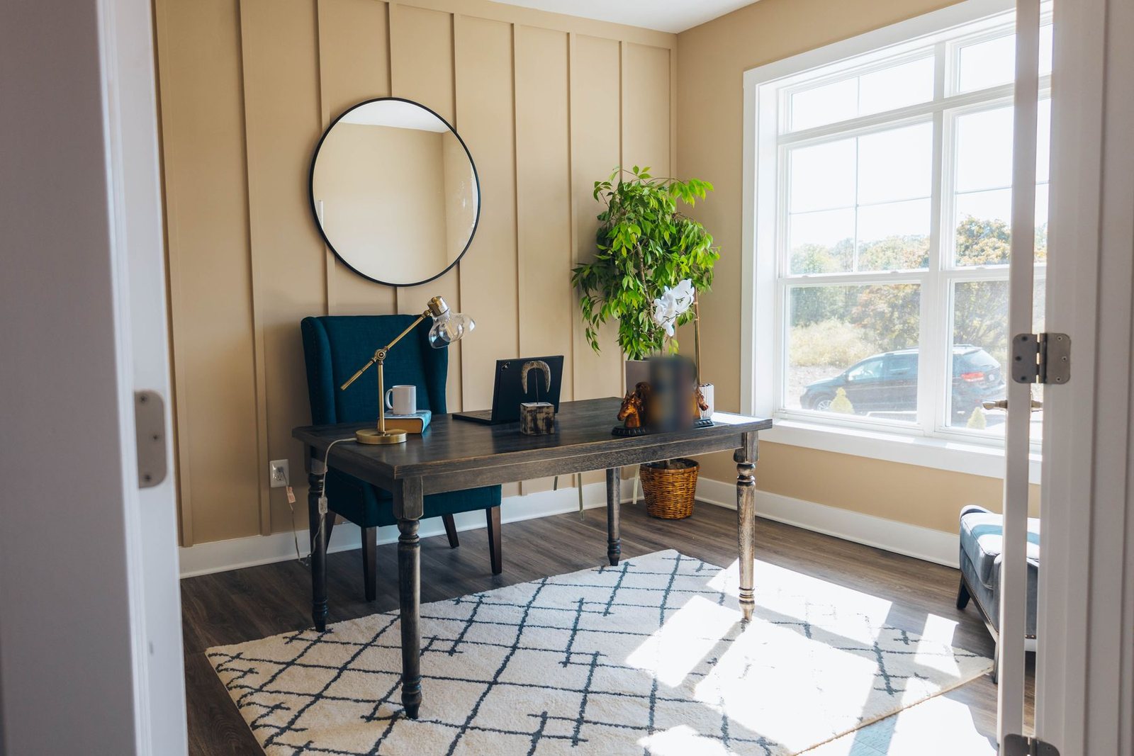 Bright home office with a dark wood desk, teal wingback chair, brass task lamp, tall potted plant and large window