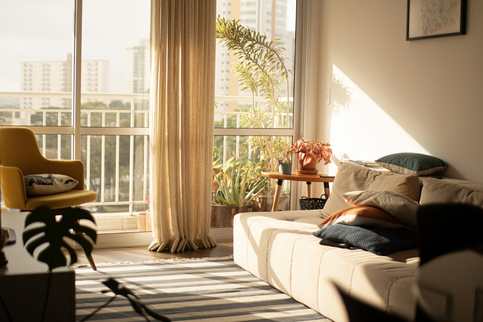 Sunlit apartment living room with linen curtains, a striped rug, potted plants, a mustard armchair and a framed print